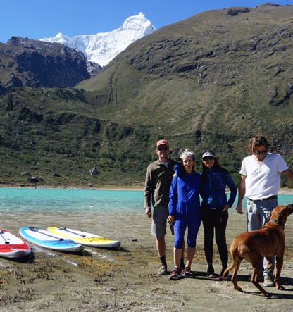 Cuatro personas de pie en la orilla de un lago con montañas y nieve en el fondo, acompañadas por un perro y varias tablas de paddle surf en la playa.