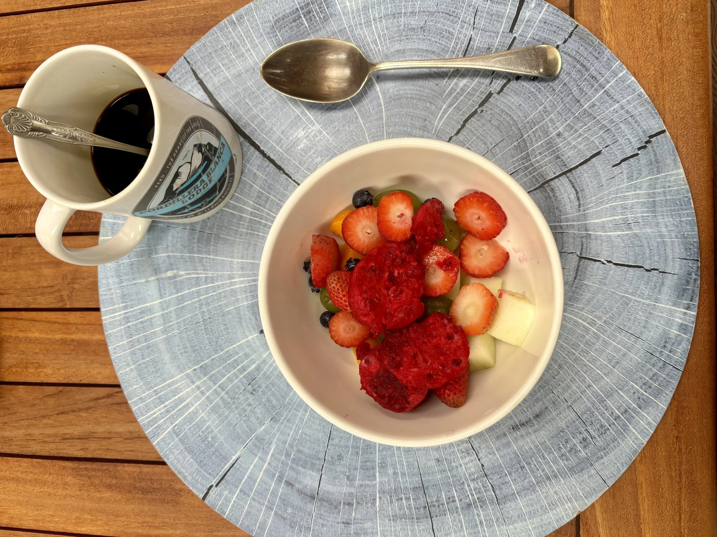 Un tazón con fruta fresca y un vaso de café negro con hielo en un plato azul y una cuchara de metal, en una mesa de madera.