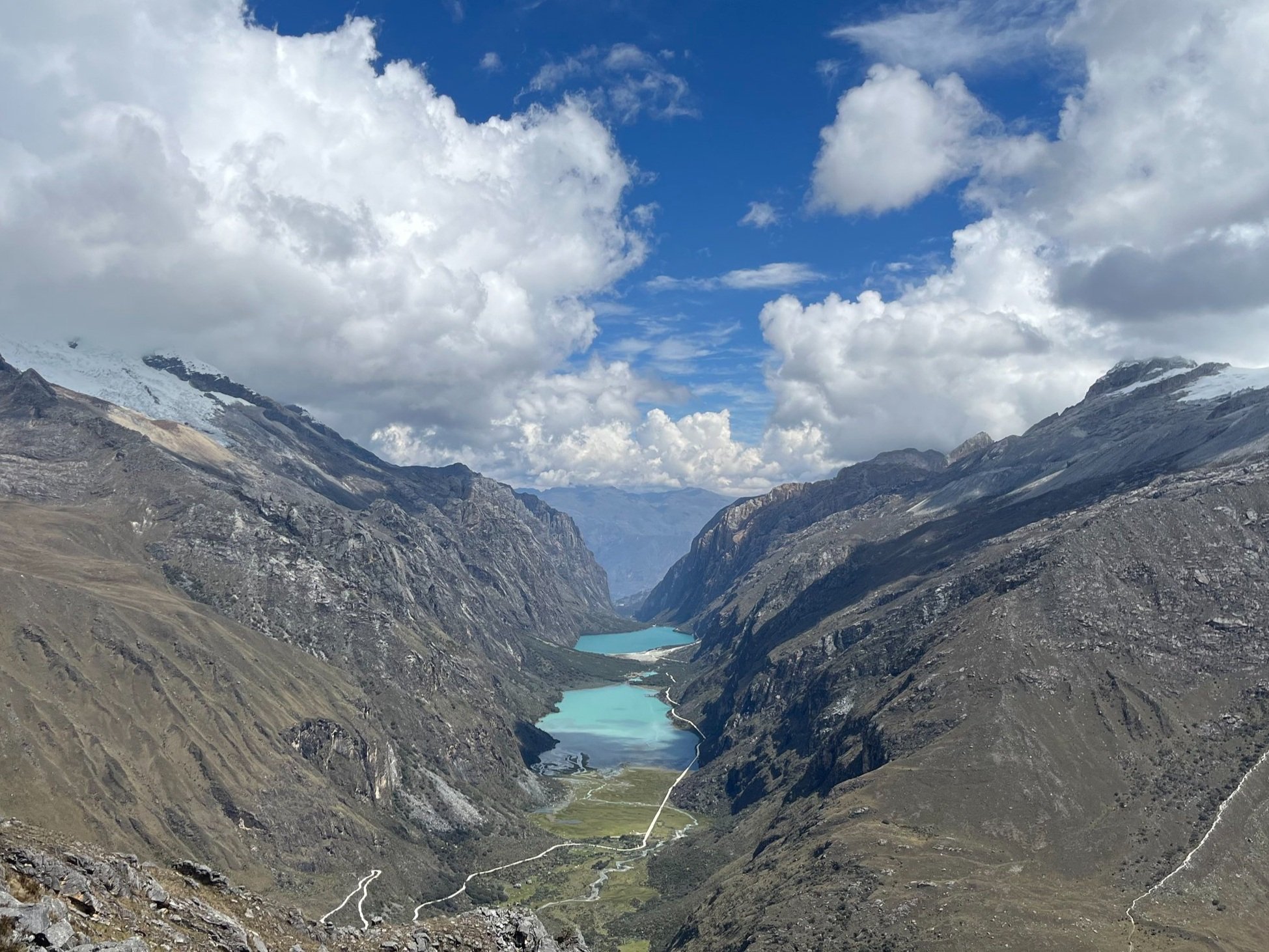 Paisaje montañoso con lagunas de color azul y caminos en el valle, rodeado de nubes y picos nevados.