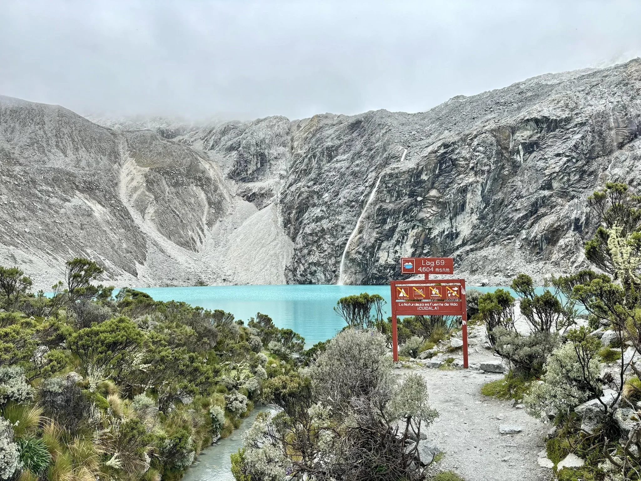 Paisaje de montañas con lago azul y vegetación en primer plano, señal que indica altura y nombre del río, y cascada en una de las montañas