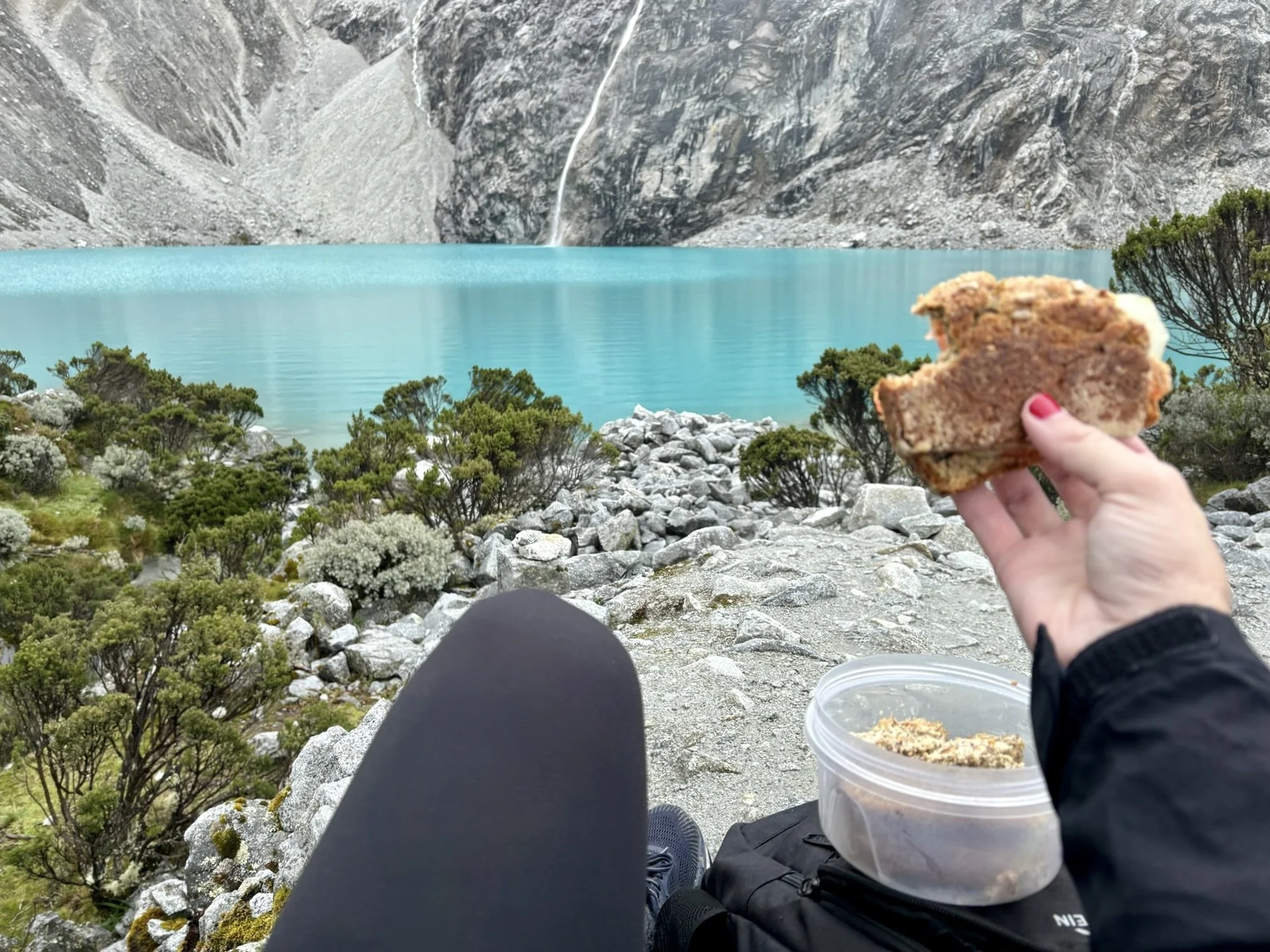 persona disfrutando de vigilando un lago de color turquesa rodeado de rocas y vegetación, sosteniendo un trozo de pastel en la mano.