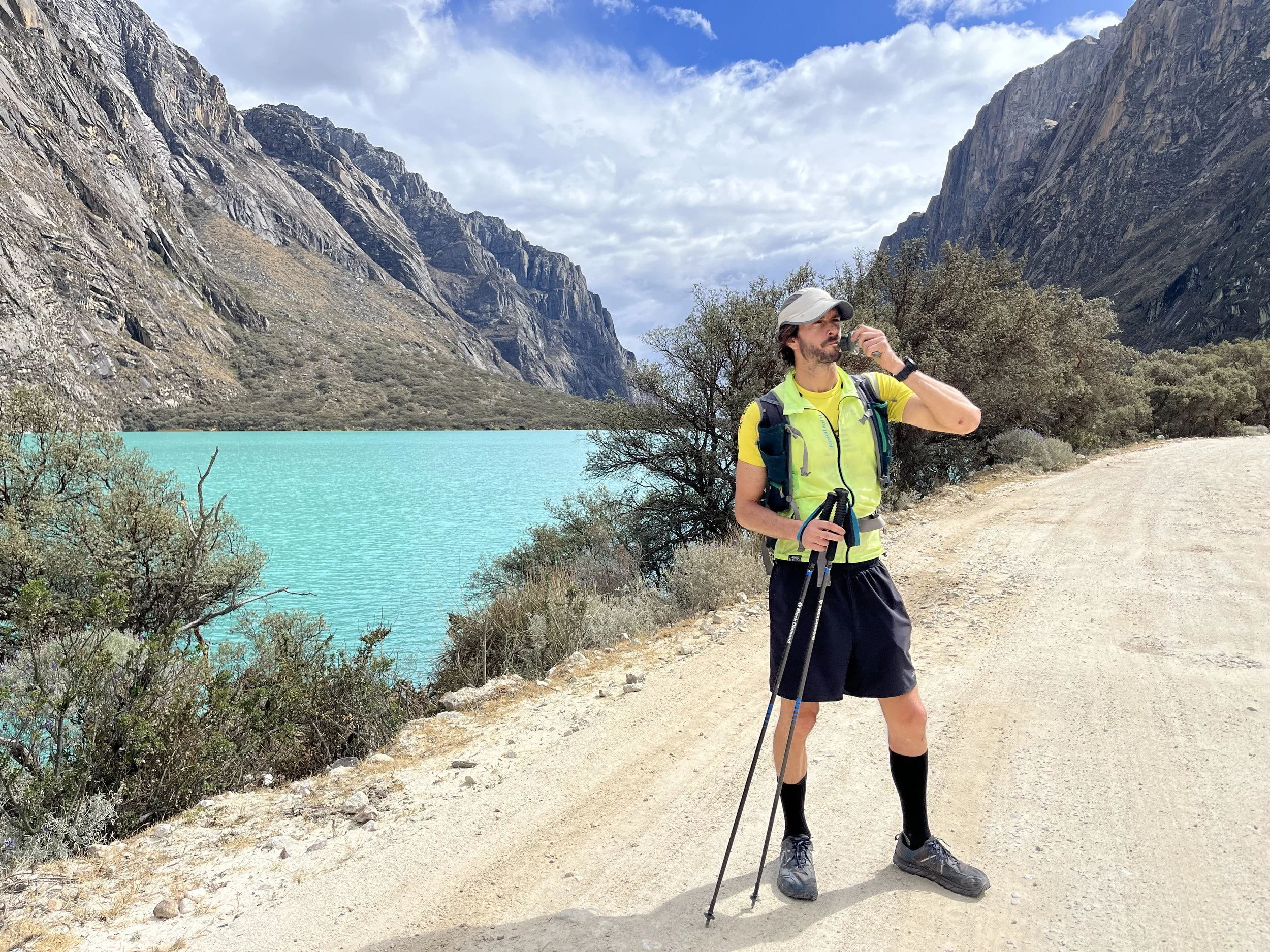 Un hombre caminando con equipo de senderismo en un paisaje con montañas, un lago de agua azul y un camino de tierra.