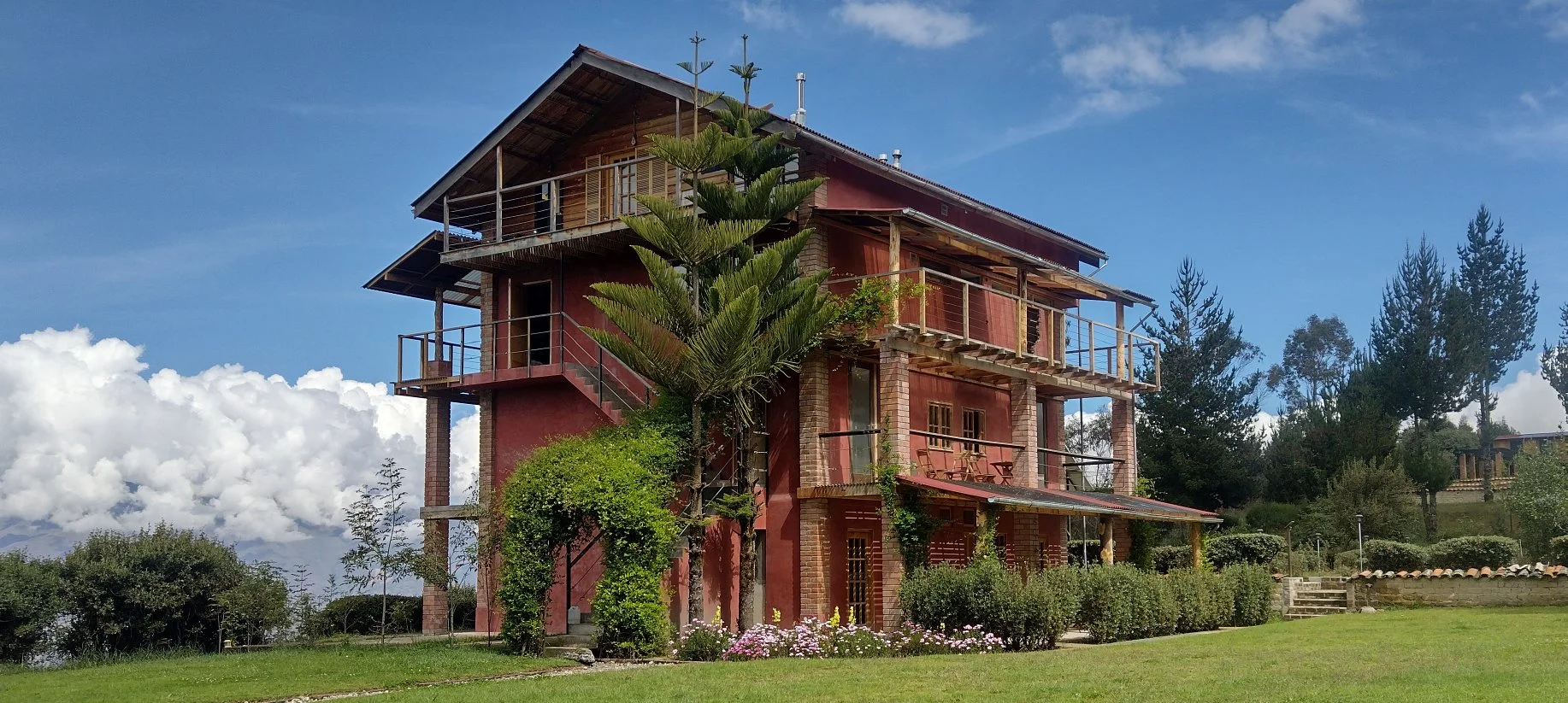 Casa de tres pisos construida con paredes de ladrillo y madera, rodeada de césped y vegetación, con un árbol grande frente a ella y un cielo azul con nubes en el fondo.
