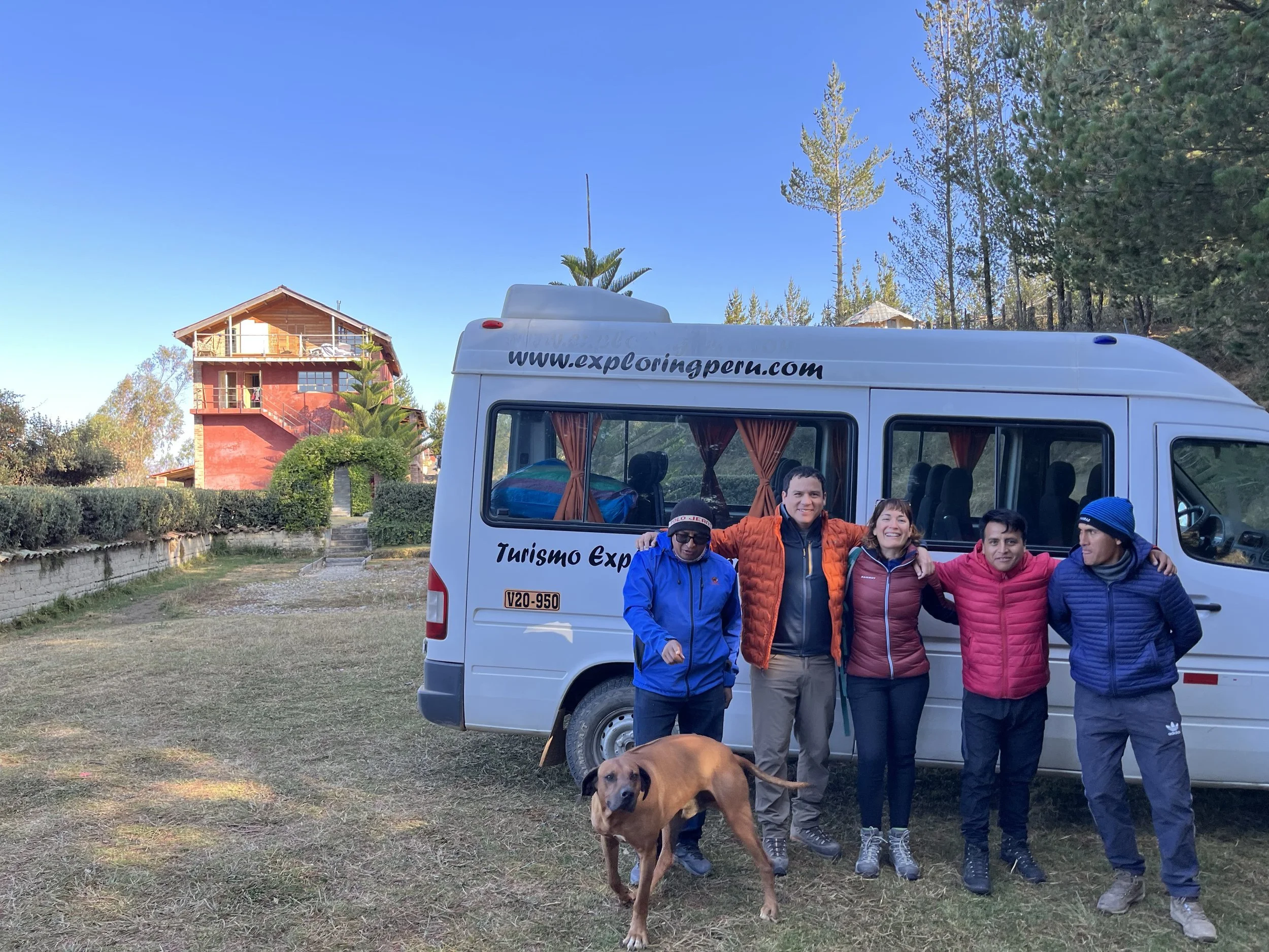 Grupo de cinco personas con chaquetas en la misma línea, frente a una camioneta blanca de turismo en un área al aire libre con árboles y una casa de madera en el fondo, acompañados por un perro marrón.