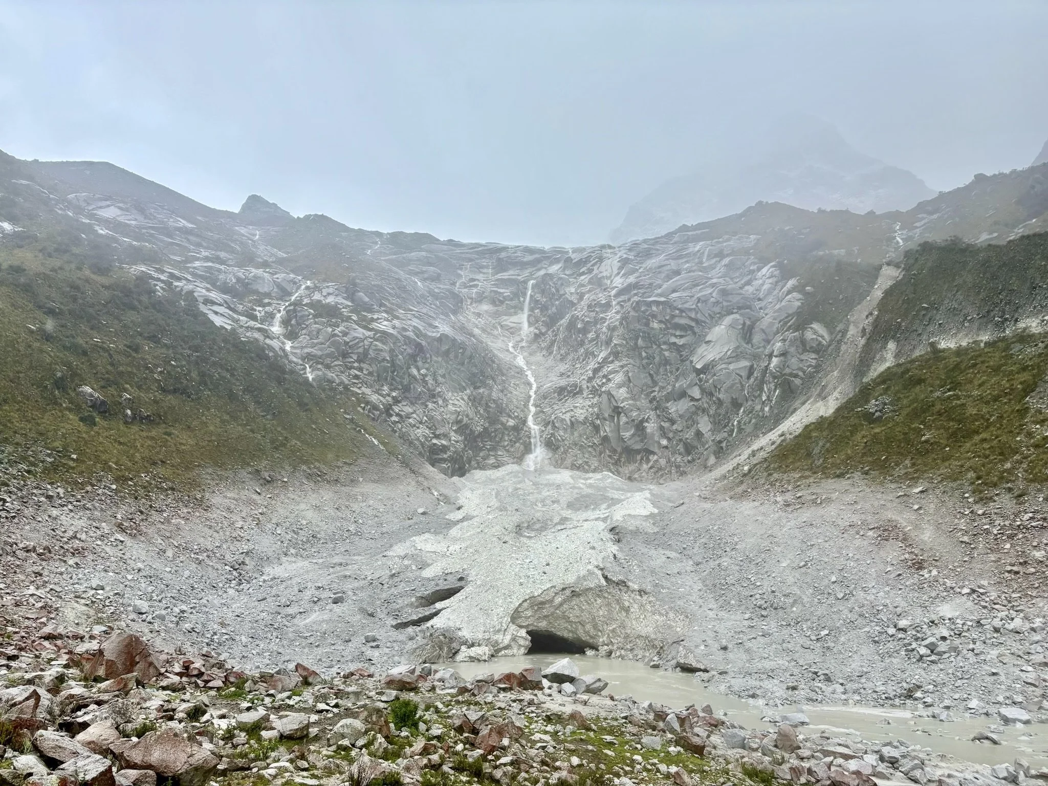 Montaña con nieve, agua de deshielo fluyendo por la roca central, suelo rocoso y vegetación dispersa.