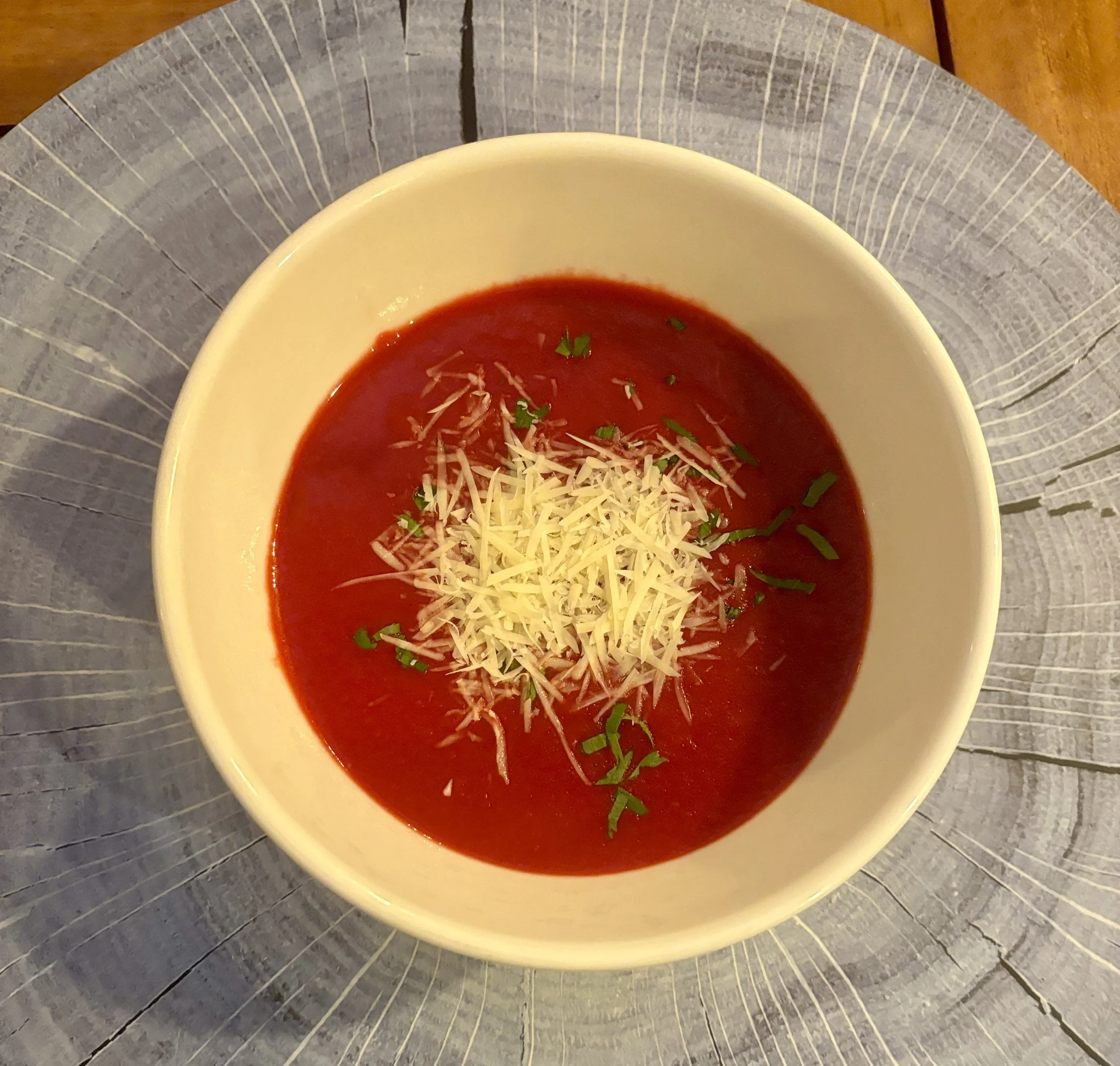 Sopa de tomate con queso rallado y perejil en un tazón blanco sobre un plato decorativo de madera.