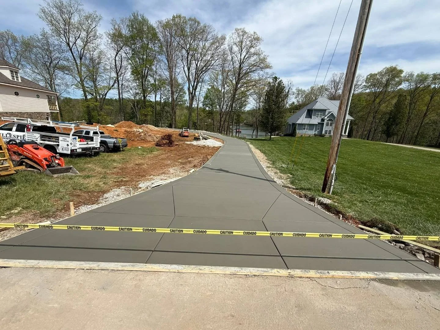 Newly poured concrete sidewalk under construction with caution tape in the foreground, construction equipment and dirt piles on the left, wooden utility poles, and houses in the background.