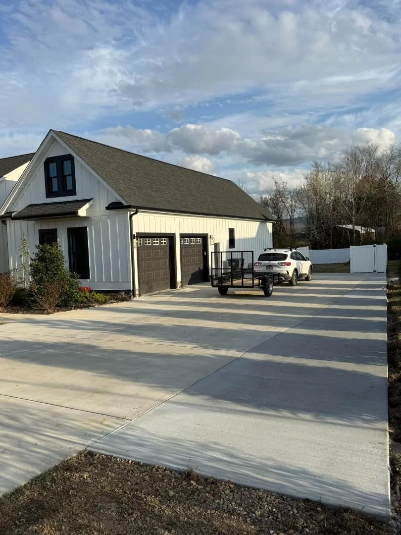 Wide concrete driveway leading to a white two-story home with dark brown double garage doors, a white SUV with a small black utility trailer parked in front