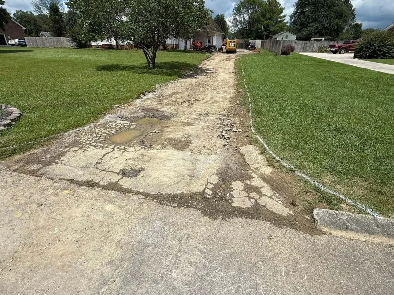 Cracked, failing residential driveway being torn out for concrete driveway replacement, with new layout marked in white spray paint and CAT skid steer on site.