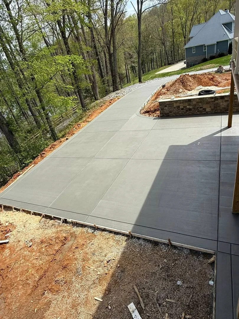 Newly poured concrete driveway leading to a house, with a wooded area on one side and a neighboring house in the background.