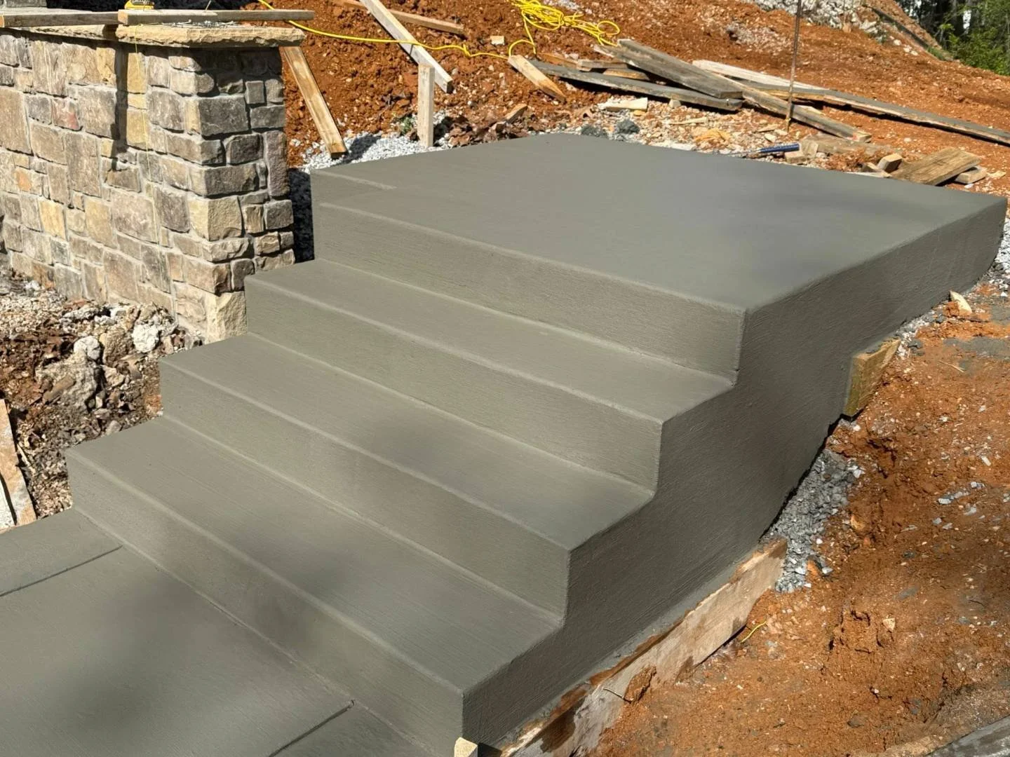 Newly poured concrete steps next to a stone wall under construction with construction materials and dirt around.