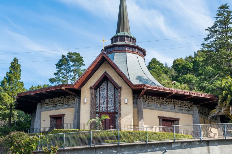 A church building with a tall steeple and cross on top, surrounded by trees, and fenced around the property.