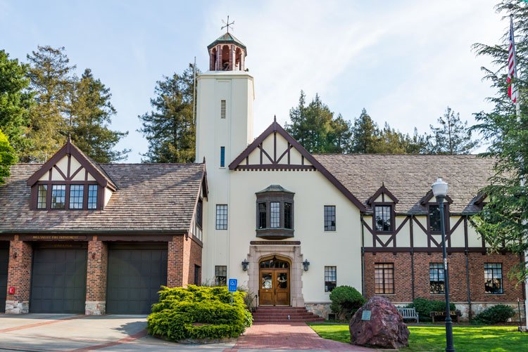 A large building resembling a house with Tudor-style architectural details, brick and stucco exterior, and a tower, surrounded by trees, with a brick pathway and green bushes in front.