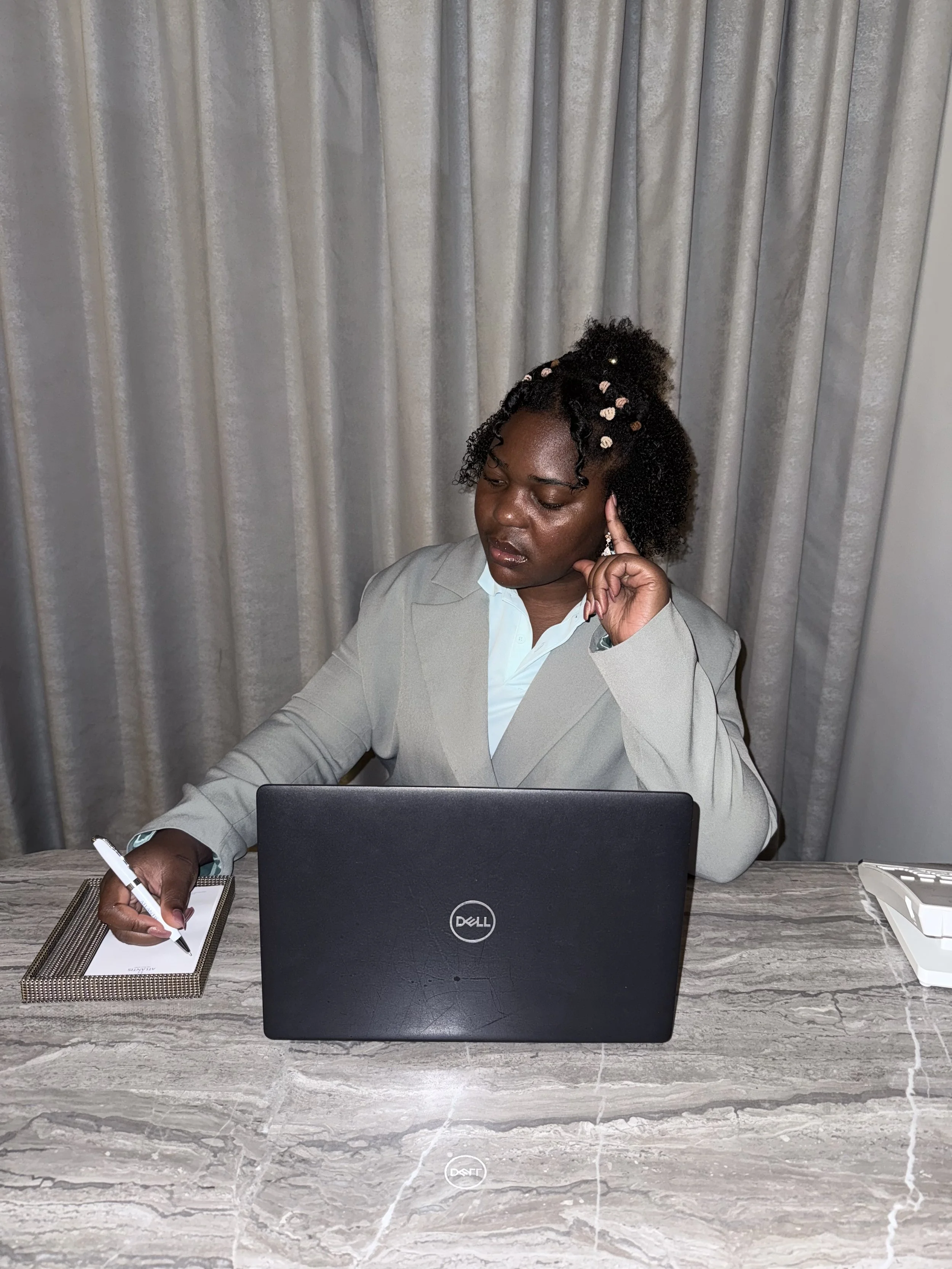 Une femme d'affaires en costume beige travaille à un bureau, écrit dans un carnet, avec un ordinateur portable Dell devant elle et un téléphone fixe à droite, avec des rideaux gris en arrière-plan.