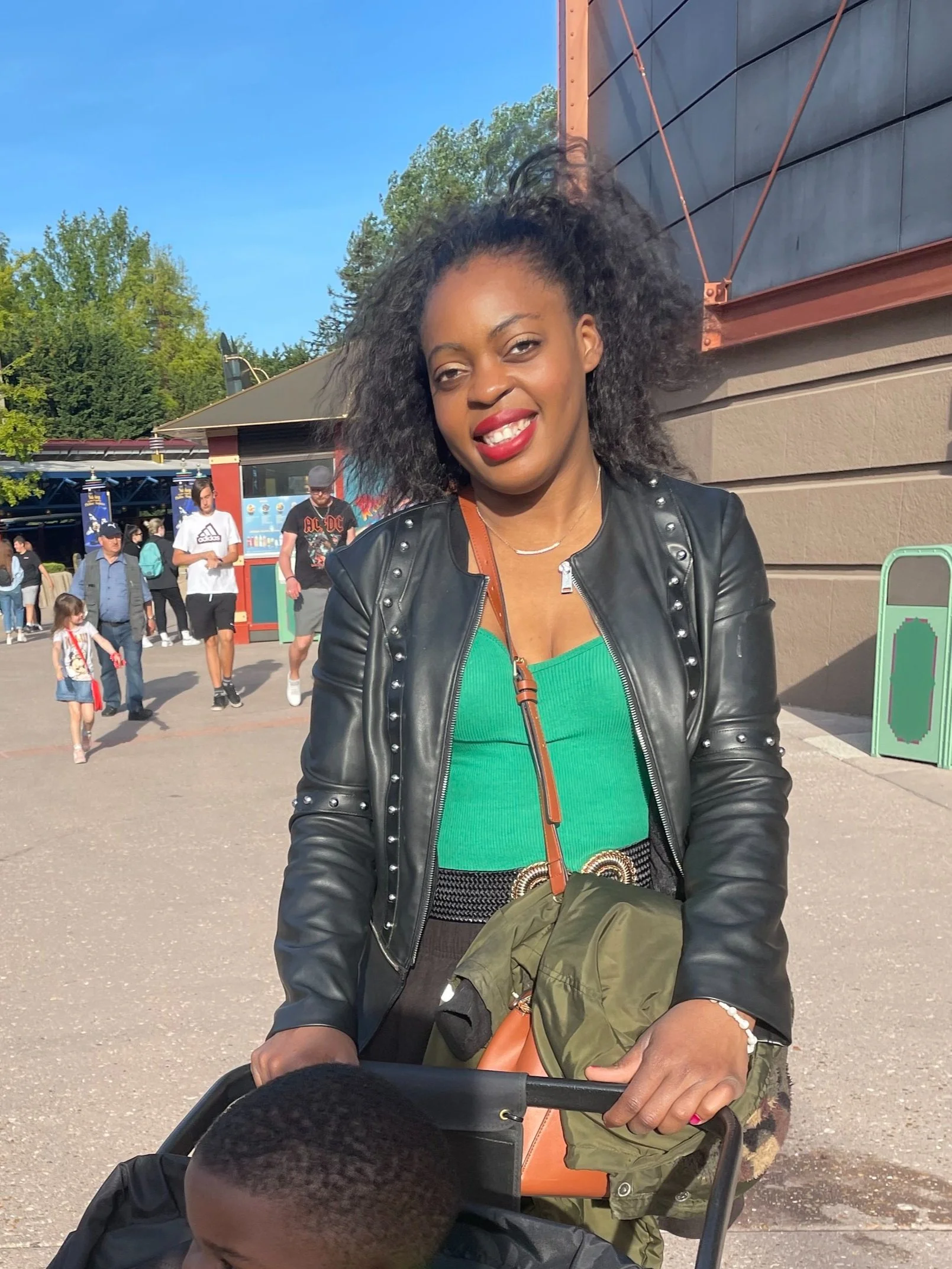 Une femme souriante portant une veste en cuir à clous, une robe verte et une ceinture à boucle, poussant une poussette, à l'extérieur par une journée ensoleillée.