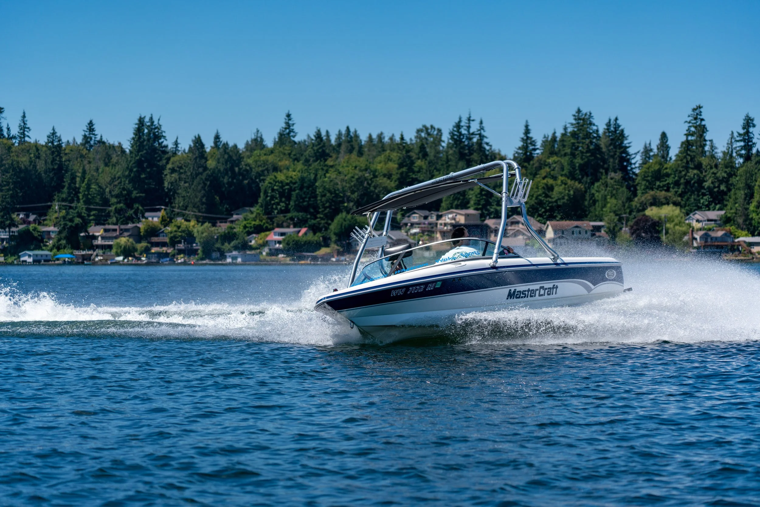 A motorboat speeding across a lake with two people inside, with houses and trees in the background.