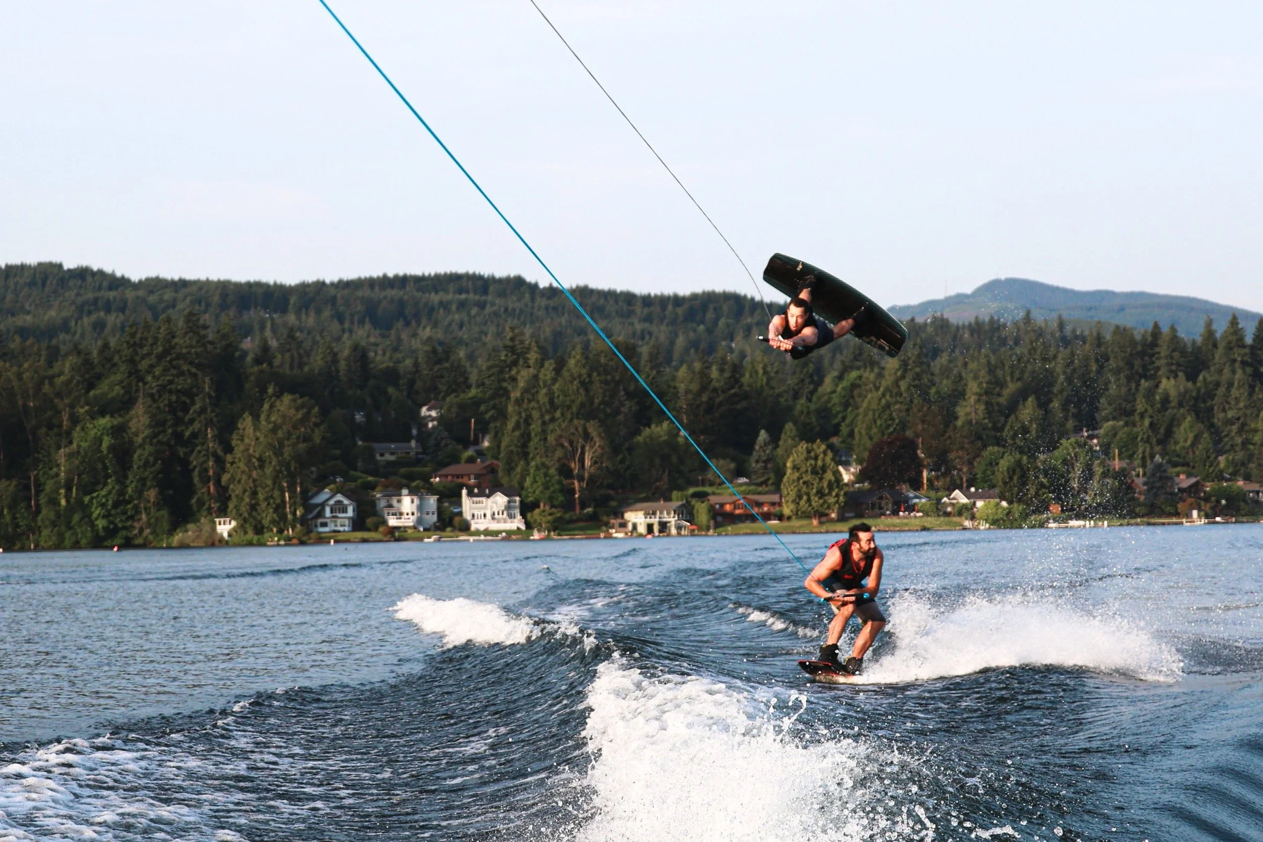 Two men wakeboarding on a lake, with one performing a jump while the other holds onto a tow rope, against a background of houses and forested hills.