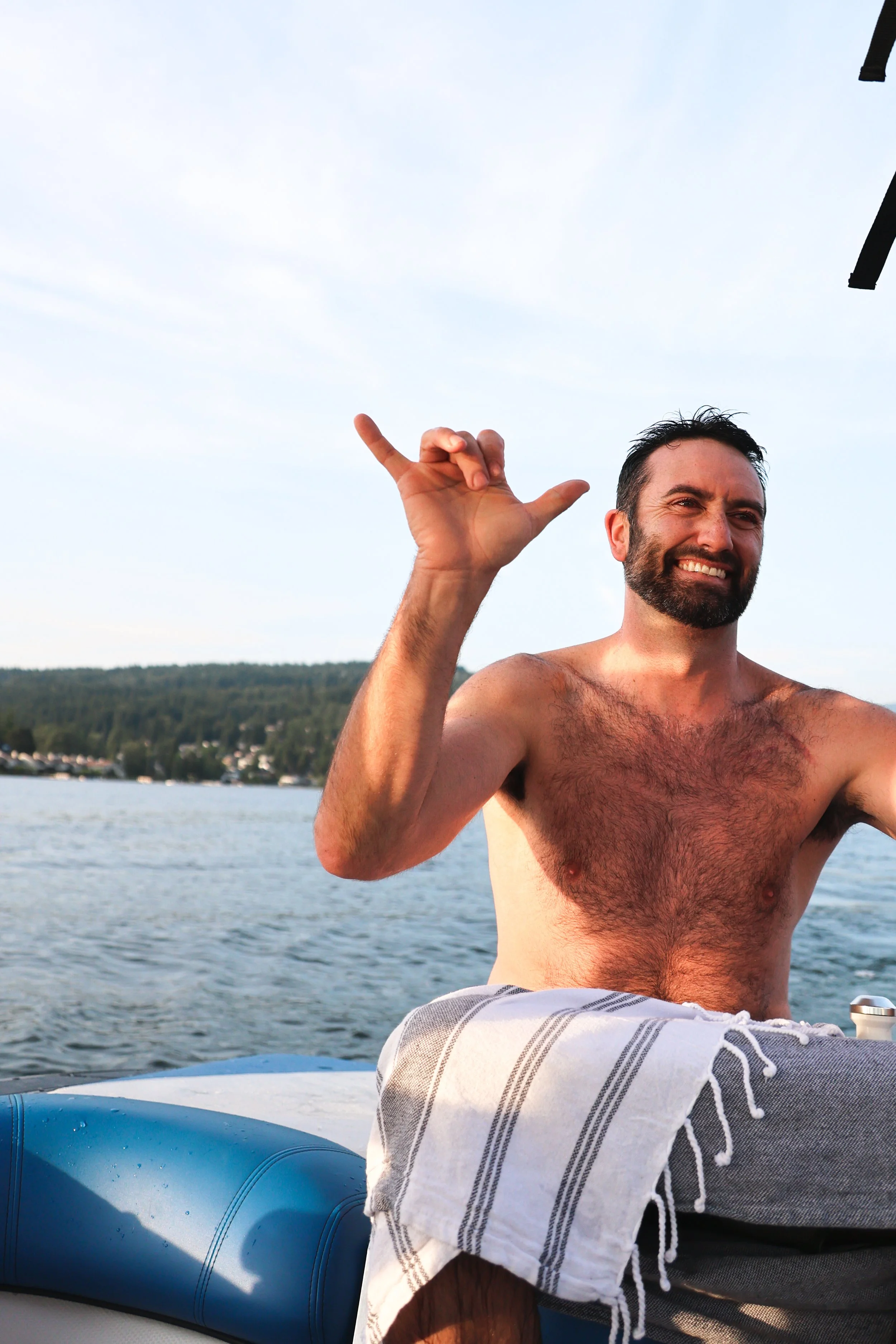 A man with a beard smiling and making a shaka sign with his hand while sitting on a boat near a body of water, with a towel draped over his lap and a scenic shoreline in the background.