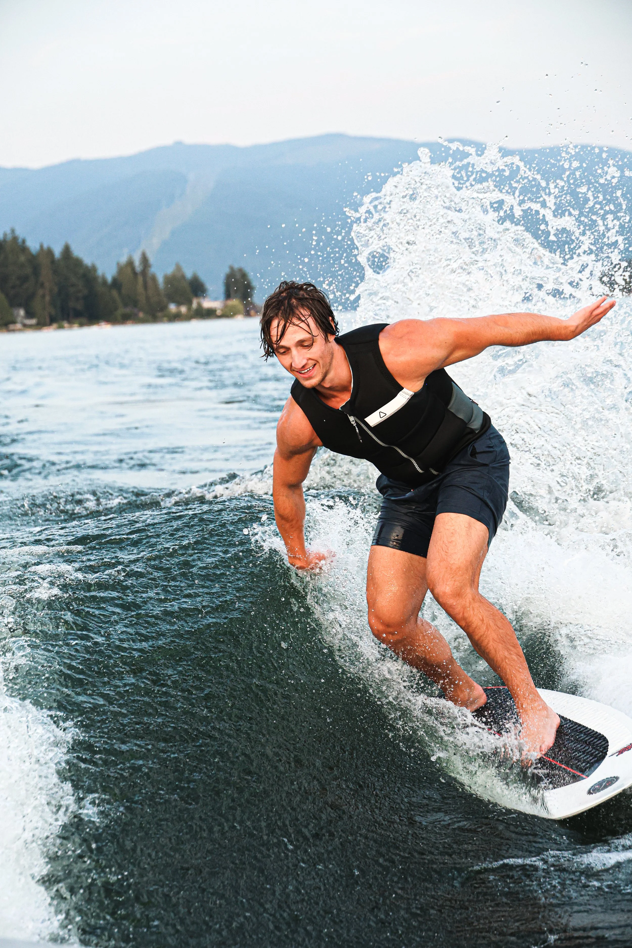 A man wakeboarding on a lake, smiling, wearing a life vest, with mountains and trees in the background.