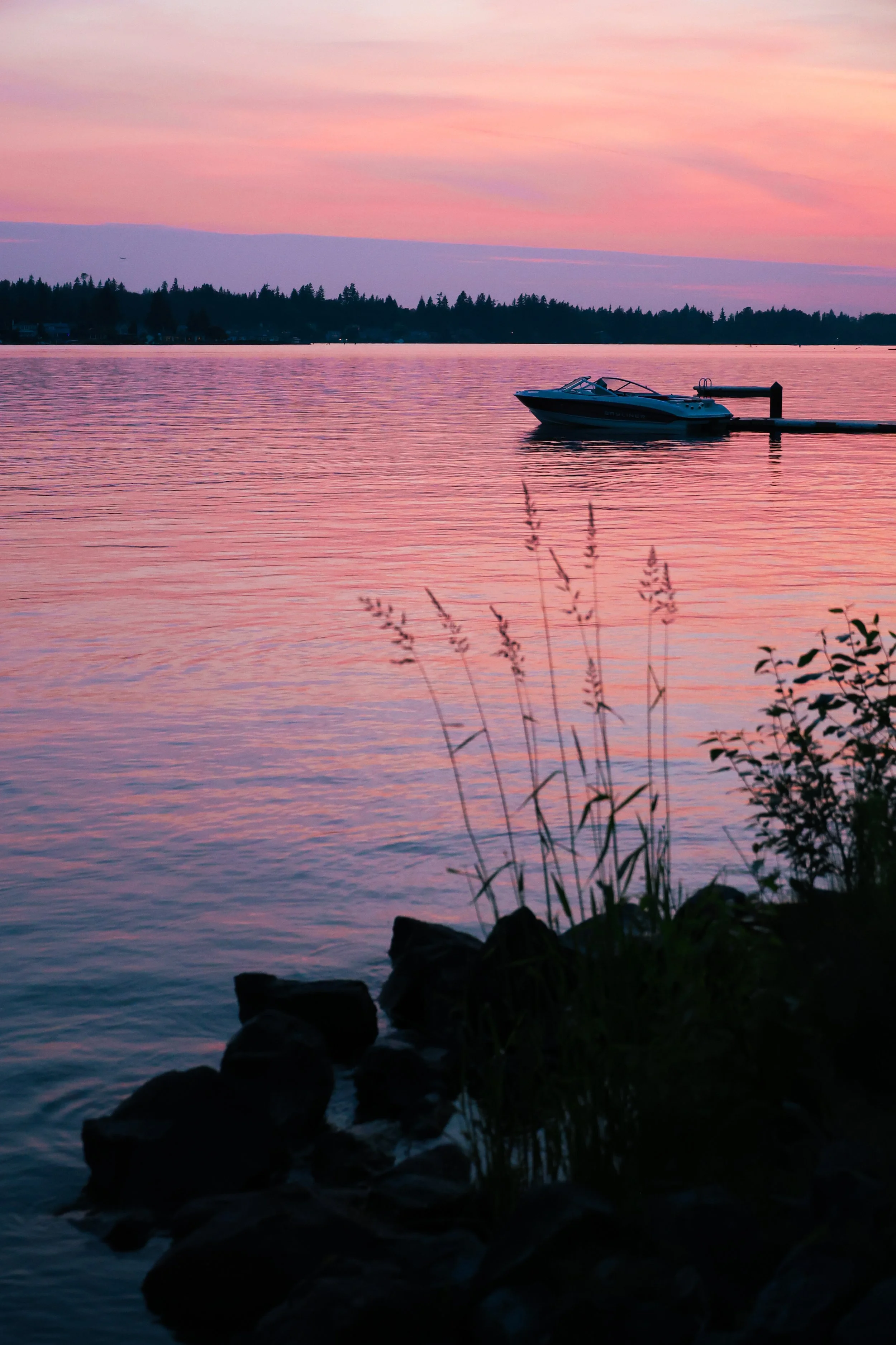 A boat docked at a pier on a calm lake during sunset with pink and purple hues in the sky and reflections on the water.