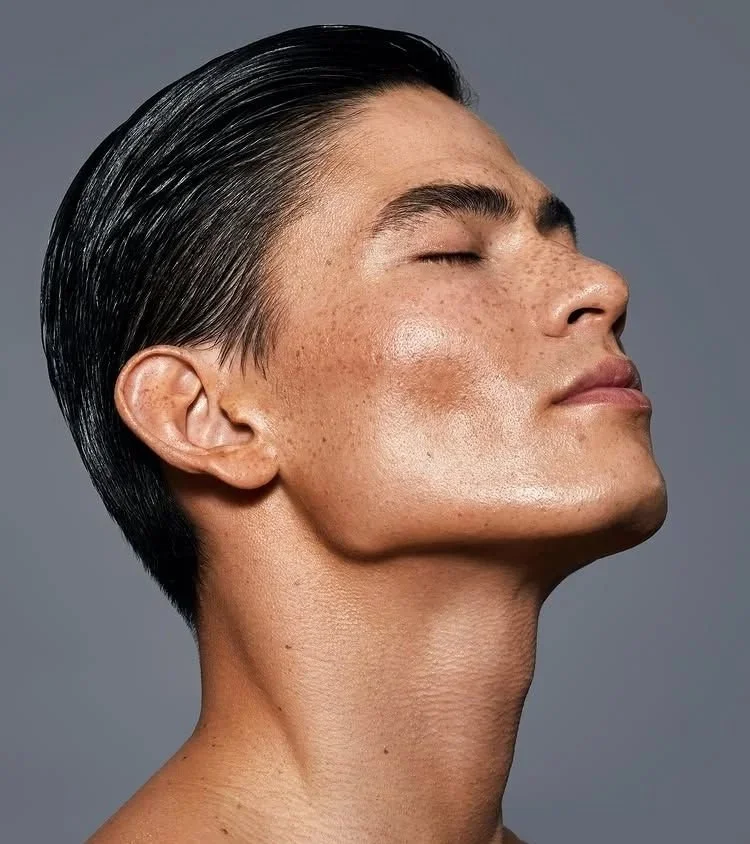 Close-up of a person with closed eyes, slicked-back hair, and glowing skin against a gray background.
