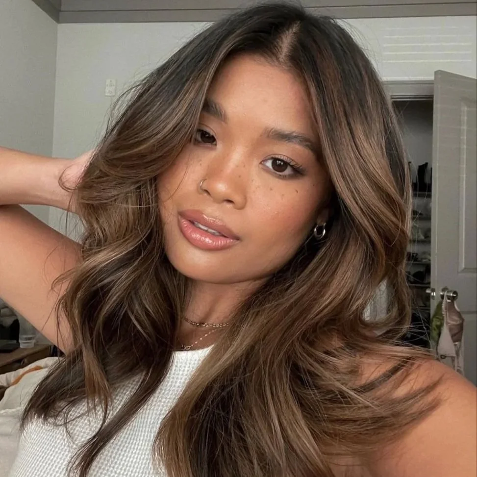 A young woman with long, wavy brown hair, wearing hoop earrings, a nose ring, and a white top, taking a selfie in a bedroom.