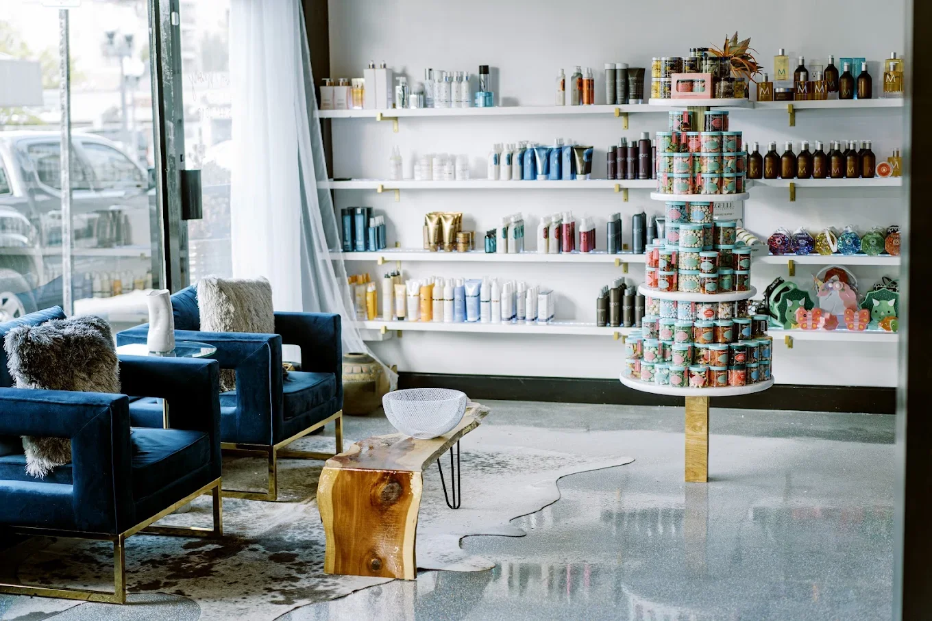 Interior of a retail store with shelves of beauty or skincare products, a display of stacked colorful tins, seating area with blue velvet chairs, a wooden coffee table, window with curtains, and decorative items.