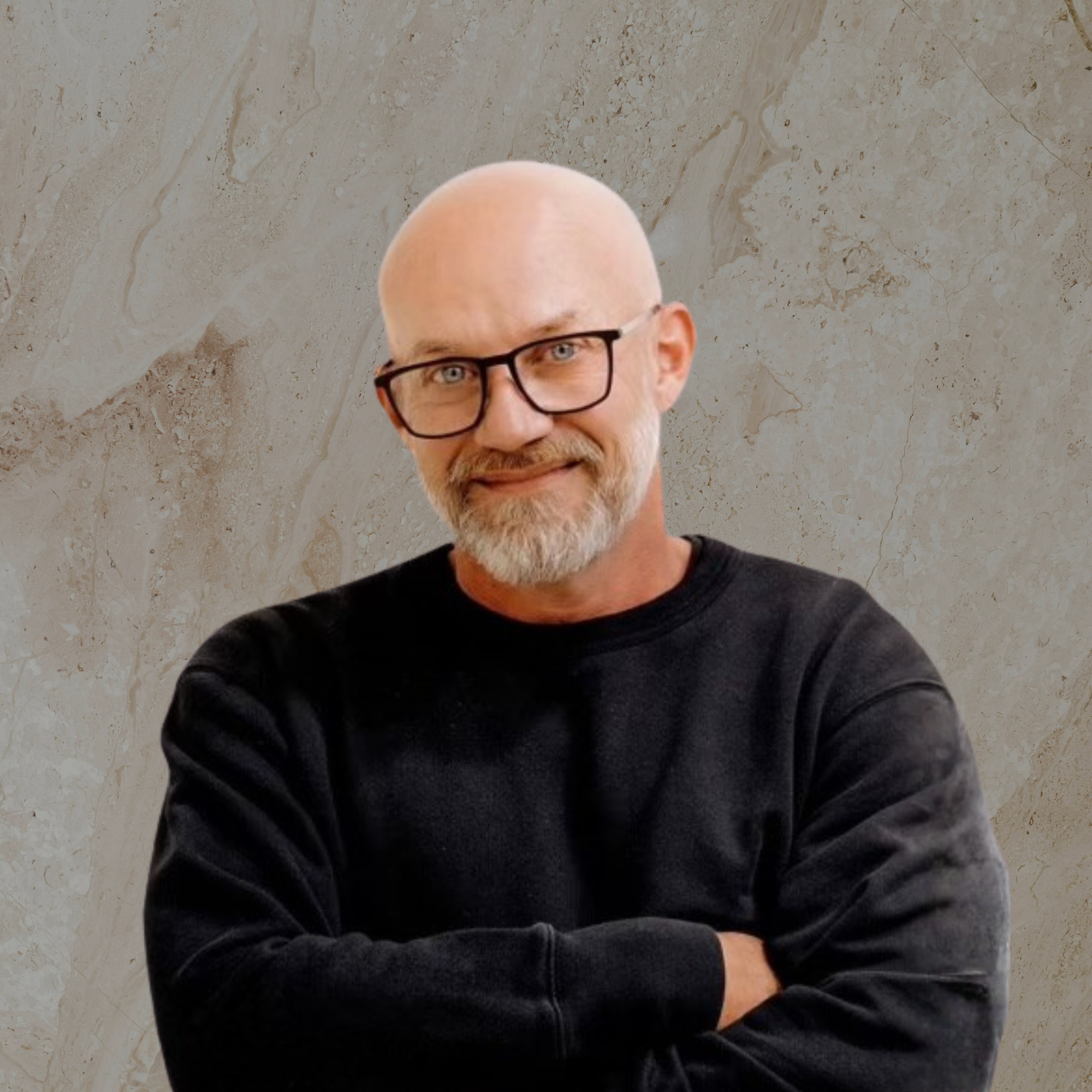 Close-up of a man with a bald head, beard, glasses, and a black shirt, smiling with folded arms, against a marble wall background.
