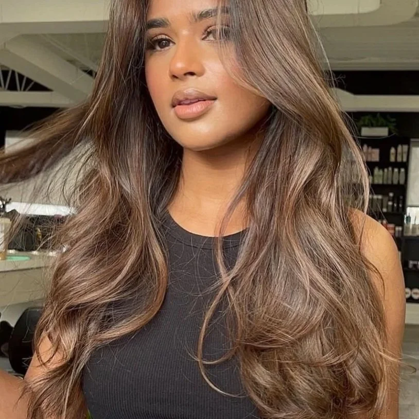 Close-up of a woman with long, wavy brown hair and light makeup, wearing a black sleeveless top, looking at the camera with a neutral expression in a salon setting.