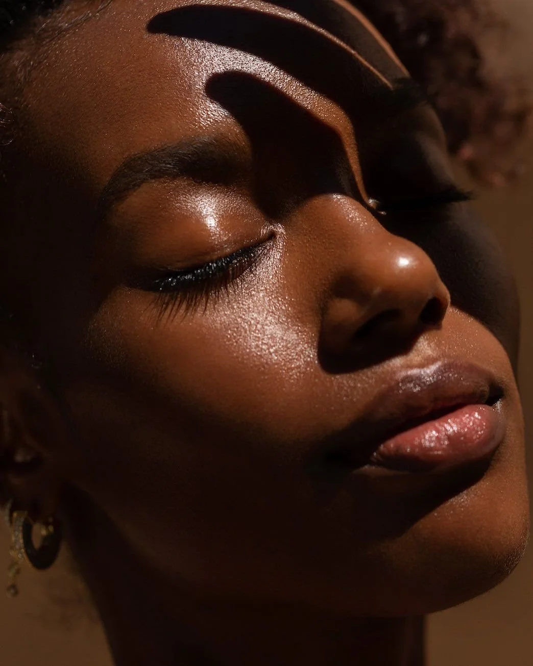 Close-up of a woman's face with her eyes closed, showing smooth, glowing skin, arched eyebrows, long eyelashes, and glossy lips, with shadows falling across her face.