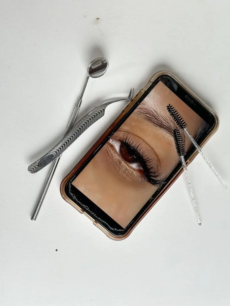 Close-up of a smartphone with an eye on the screen, surrounded by eyelash extension tools, on a white surface.