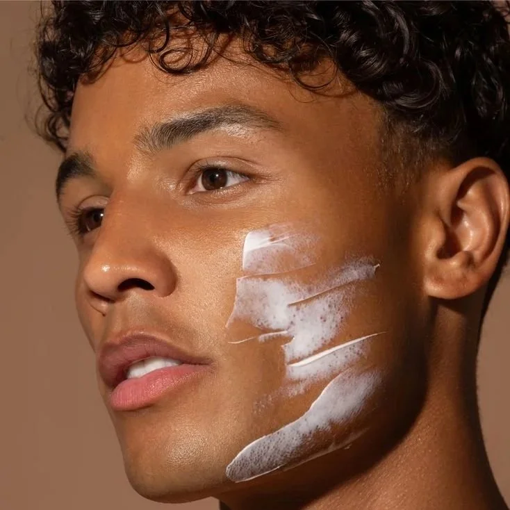 A young man with curly hair and smooth skin has white cream applied to his face.