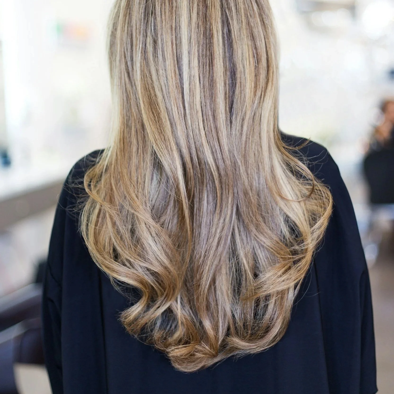Back of a person's head showing long, wavy, blonde hair with darker roots, wearing a black top, in a bright indoor setting.