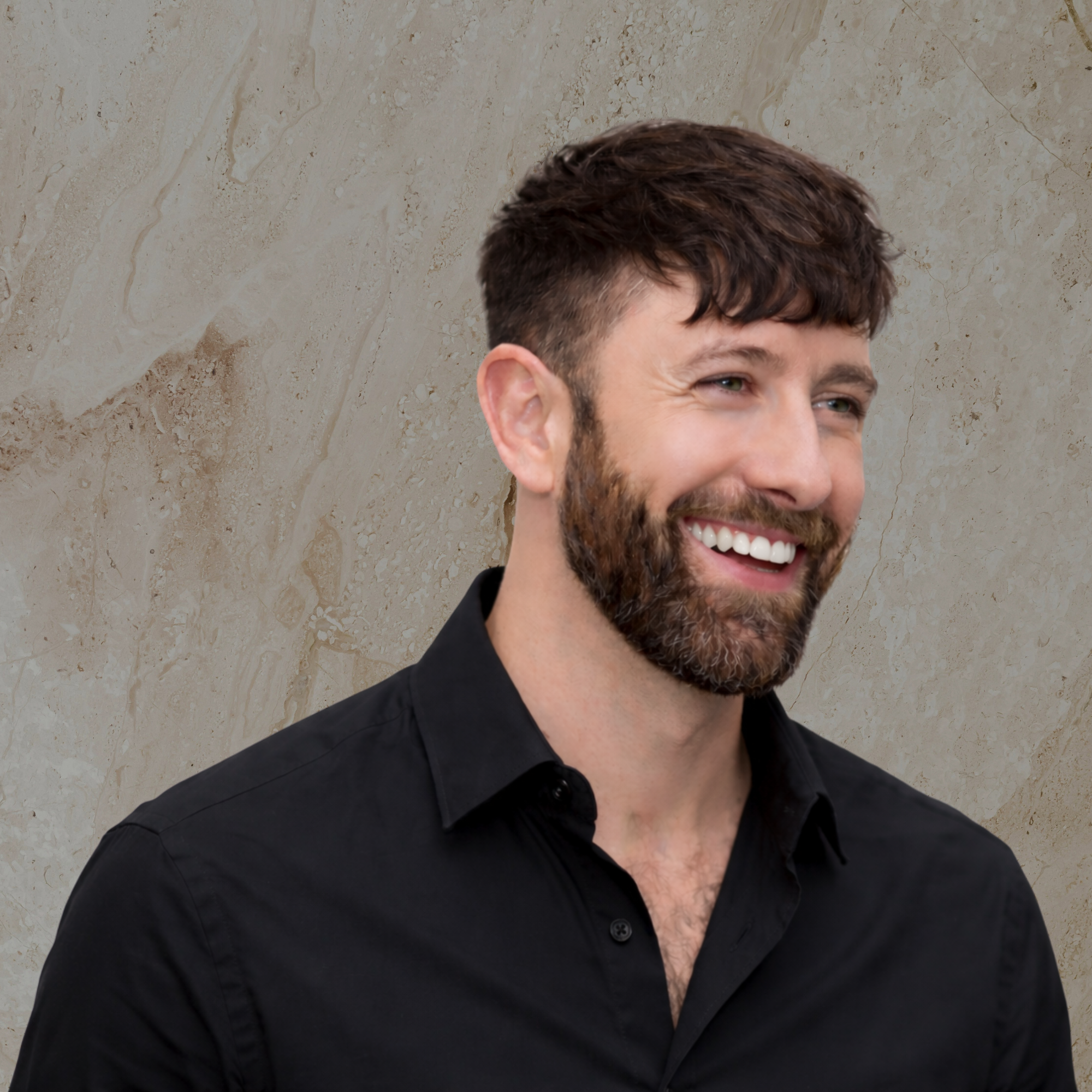 Close-up of a man with dark hair, beard, and mustache, smiling and wearing a black collared shirt, standing in front of a beige marble wall.