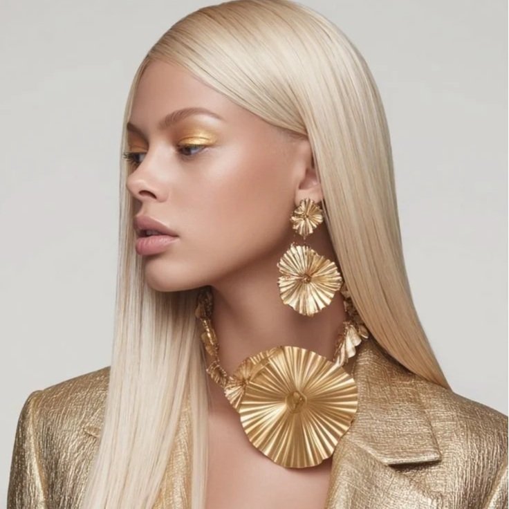 A woman with long platinum blonde hair wears bold gold earrings and a matching chunky gold necklace, and a gold metallic blazer.