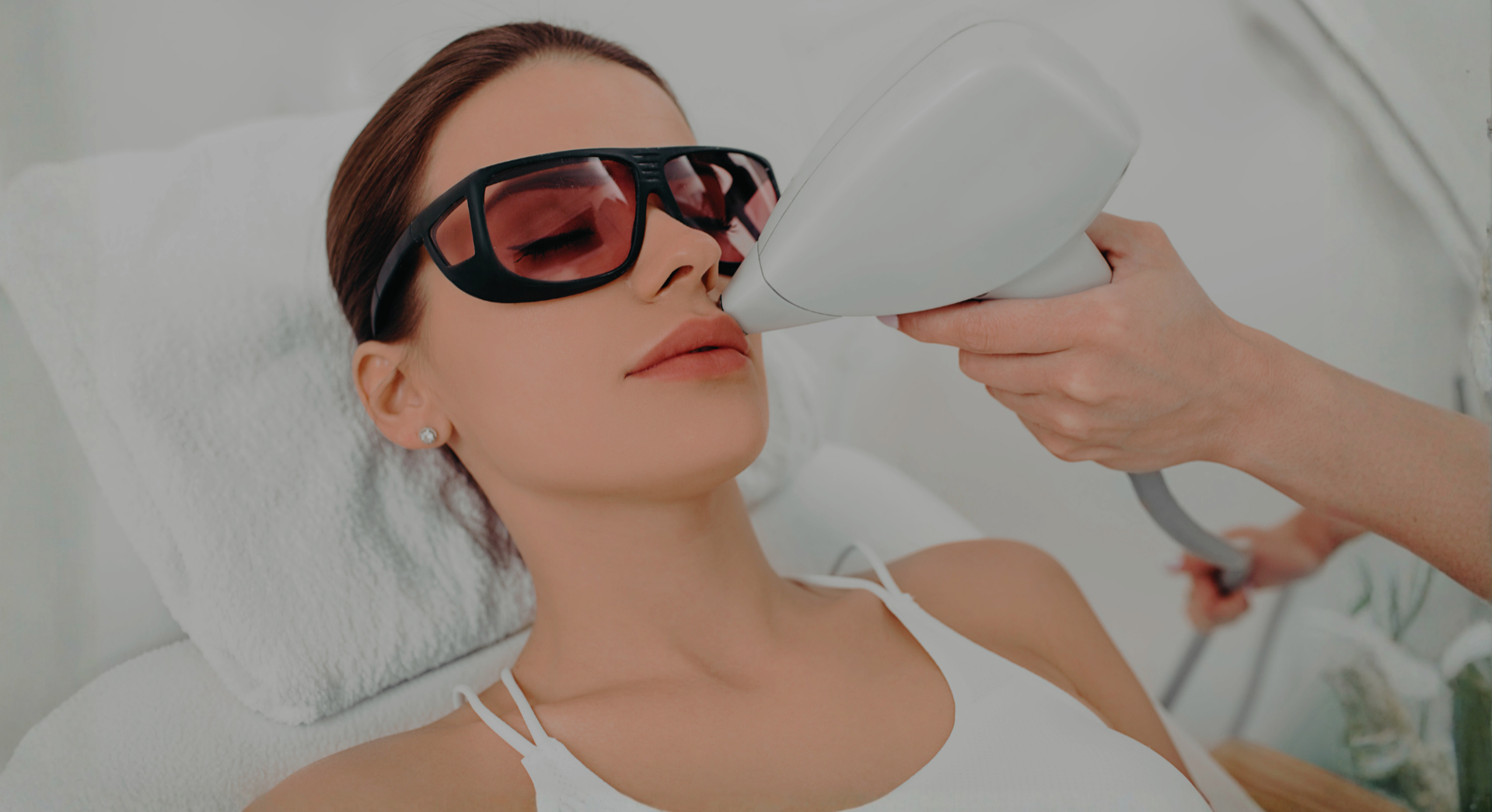 A woman lying on a medical bed wearing protective glasses while a healthcare professional uses a laser device on her face.