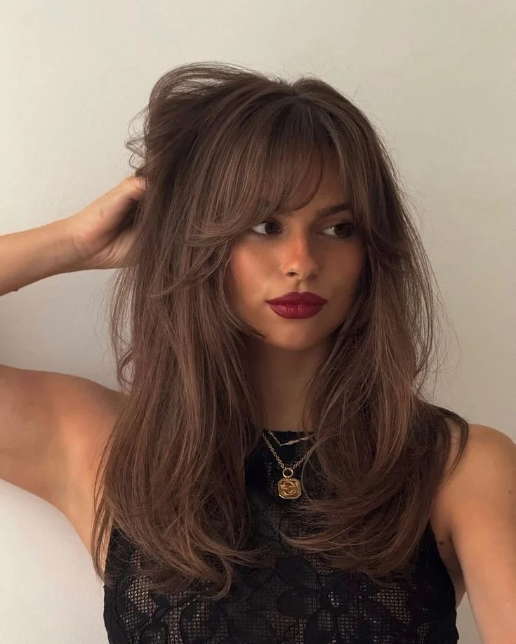 A woman with styled, voluminous brown hair, wearing a black lace top and a gold necklace with a pendant, posing against a plain background.