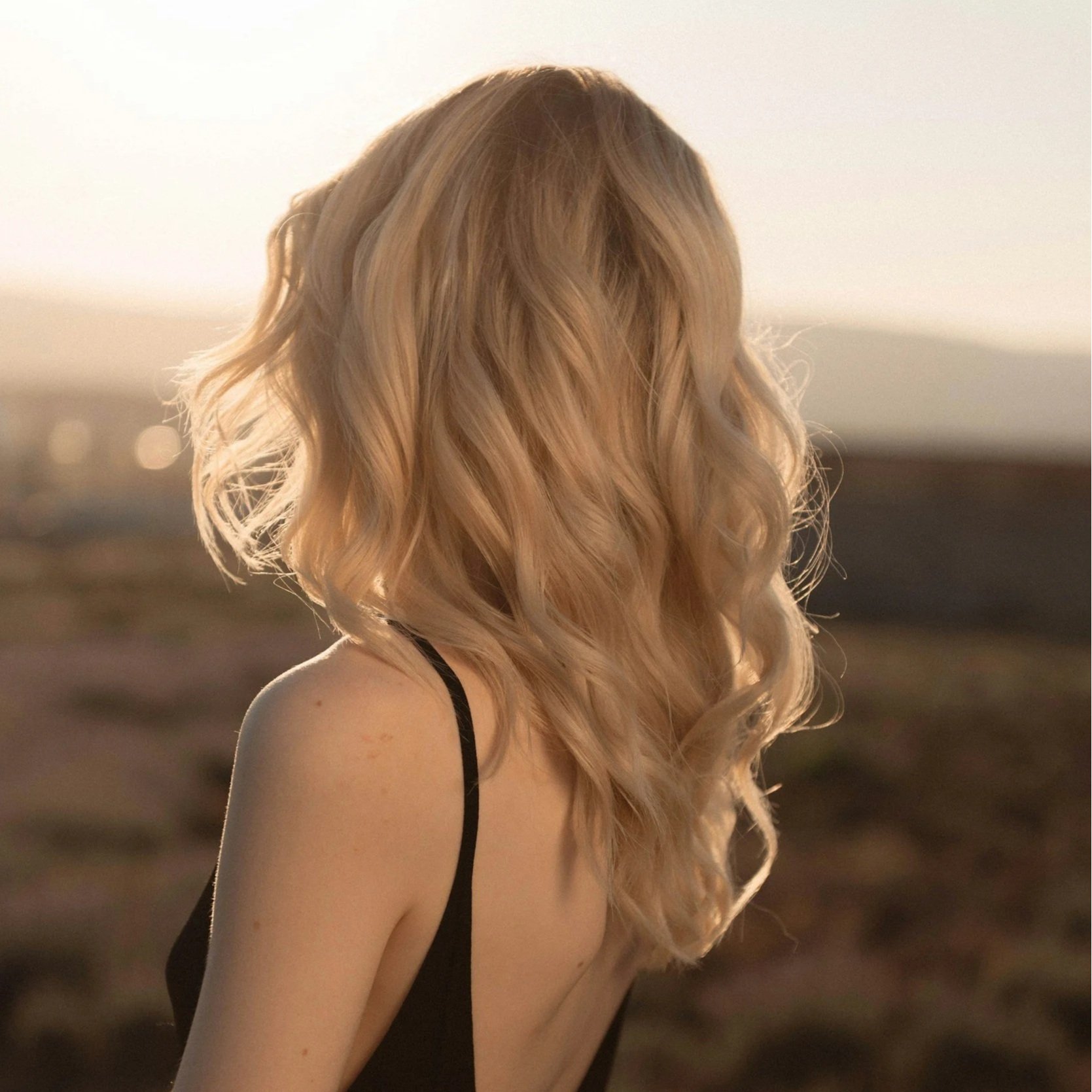 Side view of a woman with long, wavy blonde hair wearing a black tank top, standing outdoors during sunset or dusk.
