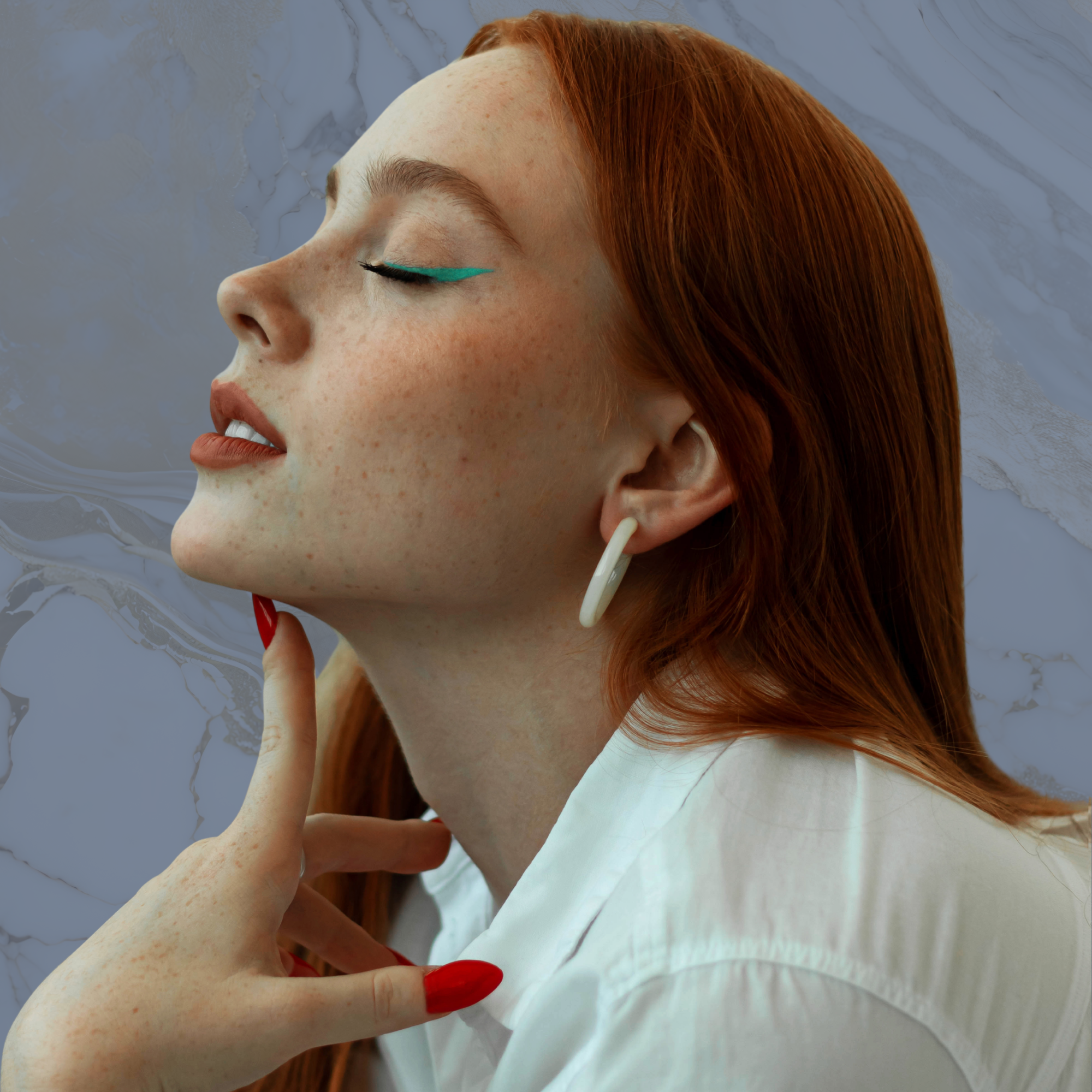Side profile of a woman with freckles, long red hair, and teal eyeliner, gently touching her chin with her hand, wearing white earrings and a white top, against a textured gray background.