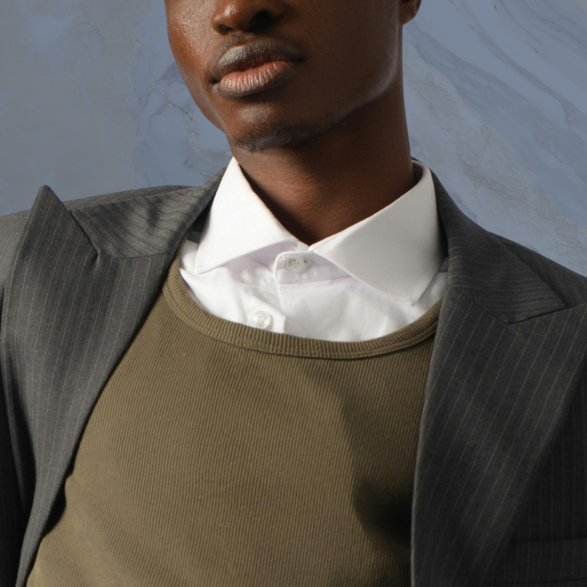 Close-up of a person with dark skin wearing a suit, white shirt, and beige sweater, with a marble textured background.