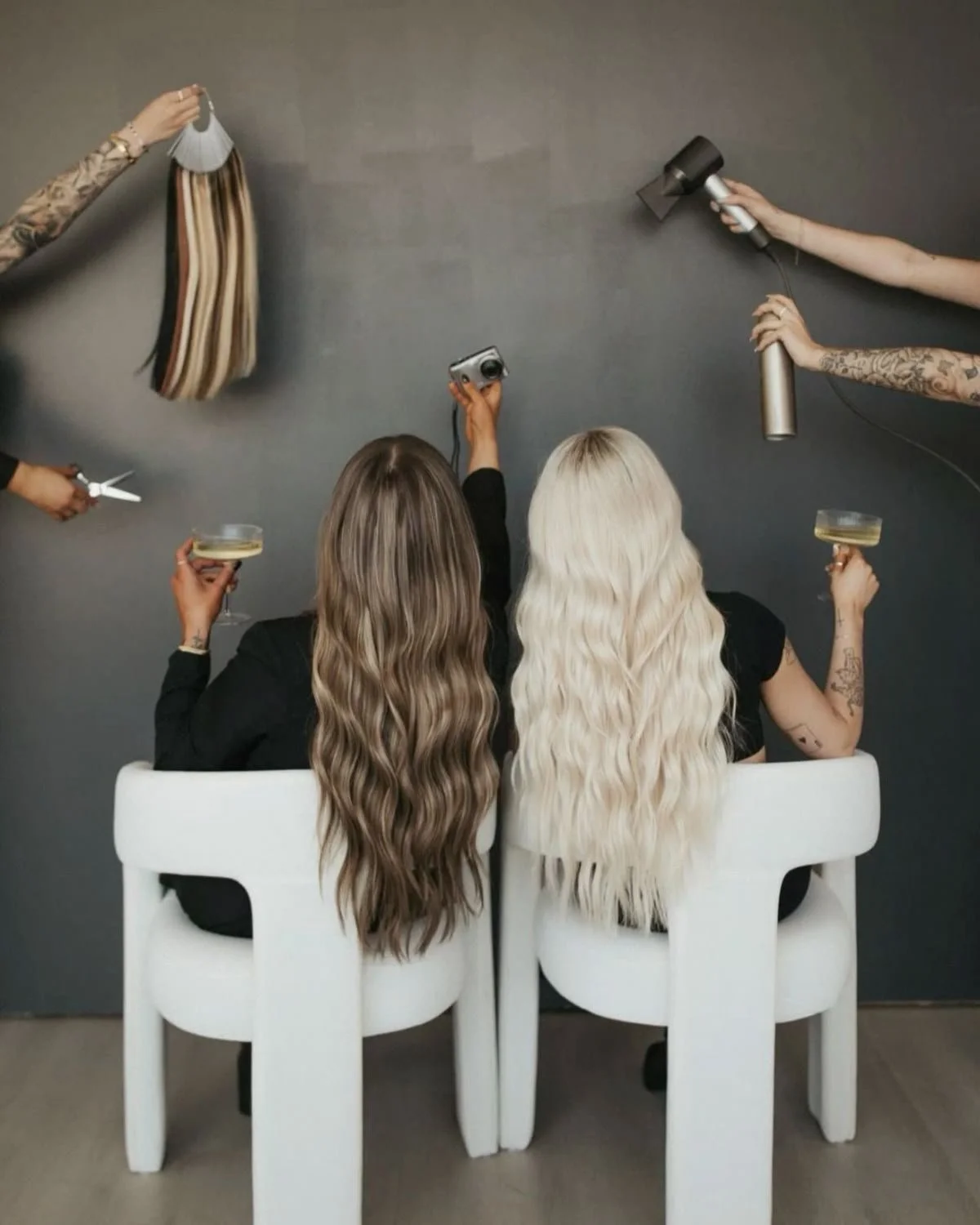Two women with long, wavy hair, one brown and one platinum blonde, sit back-to-back on white chairs, holding champagne glasses. Behind them, several hands are styling their hair with hair tools, a blow dryer, a curling iron, and a comb, against a plain gray wall.
