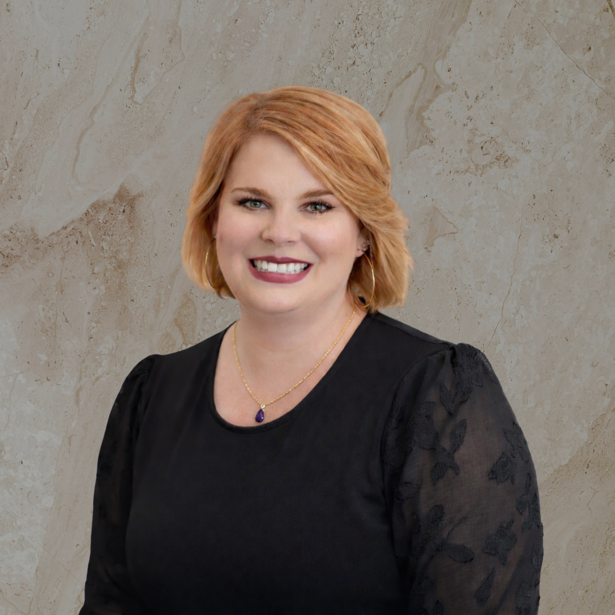 Close-up of a woman with short, wavy, blonde hair against a beige marble background. She is smiling, showing her teeth, and wearing a black top, gold earrings, a gold necklace with a purple gemstone pendant, and makeup with dark lipstick.