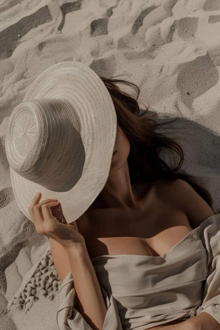 Woman lying on the sand at the beach, wearing a large sun hat and a beige off-shoulder dress.
