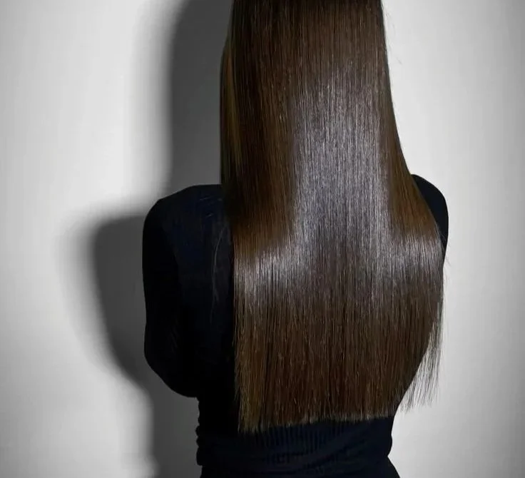 A woman with long, straight, shiny brown hair facing a white wall.