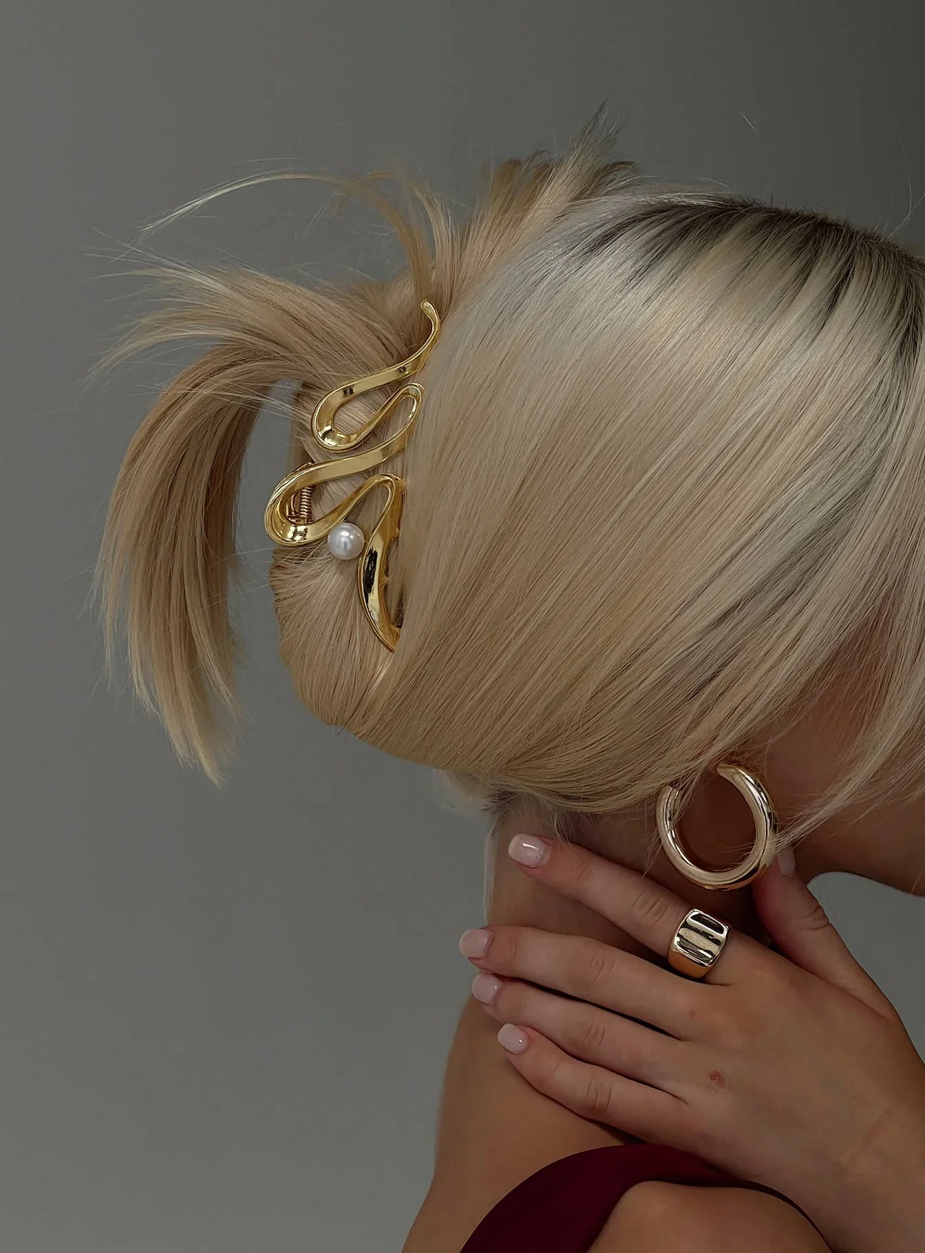 Close-up of a woman with blonde hair, styled in an elegant updo, accessorized with a gold hair clip and pearl decoration. She wears large hoop earrings and rings, with her hand resting on her shoulder.