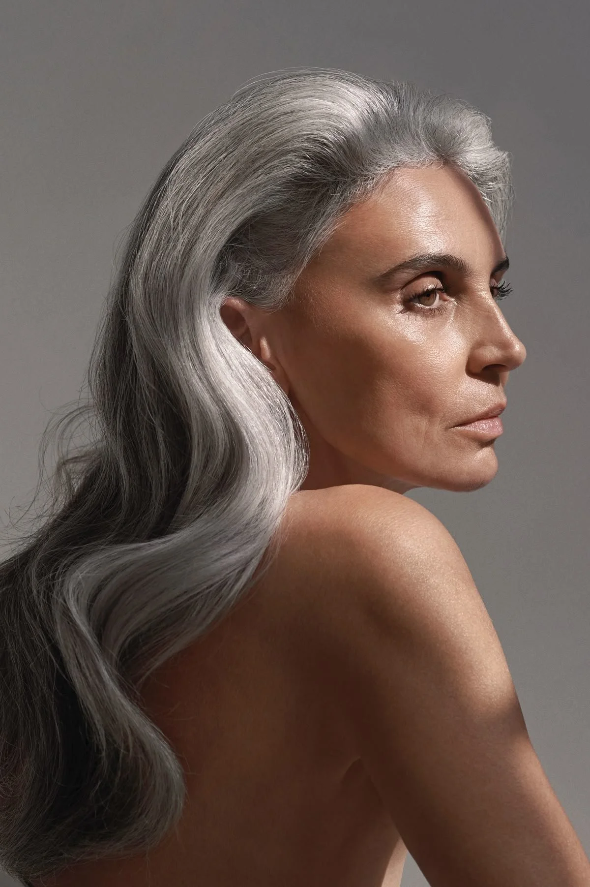 A side profile of a mature woman with long, wavy gray hair, looking pensively into the distance against a neutral gray background.