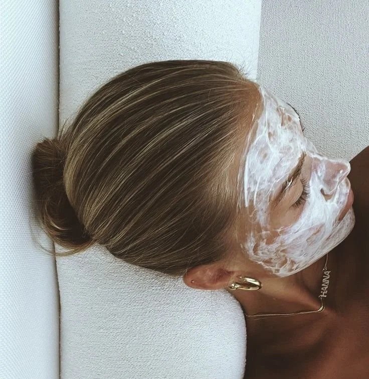 Woman lying on a white couch with a cream face mask on her face, eyes closed, and hair in a bun.