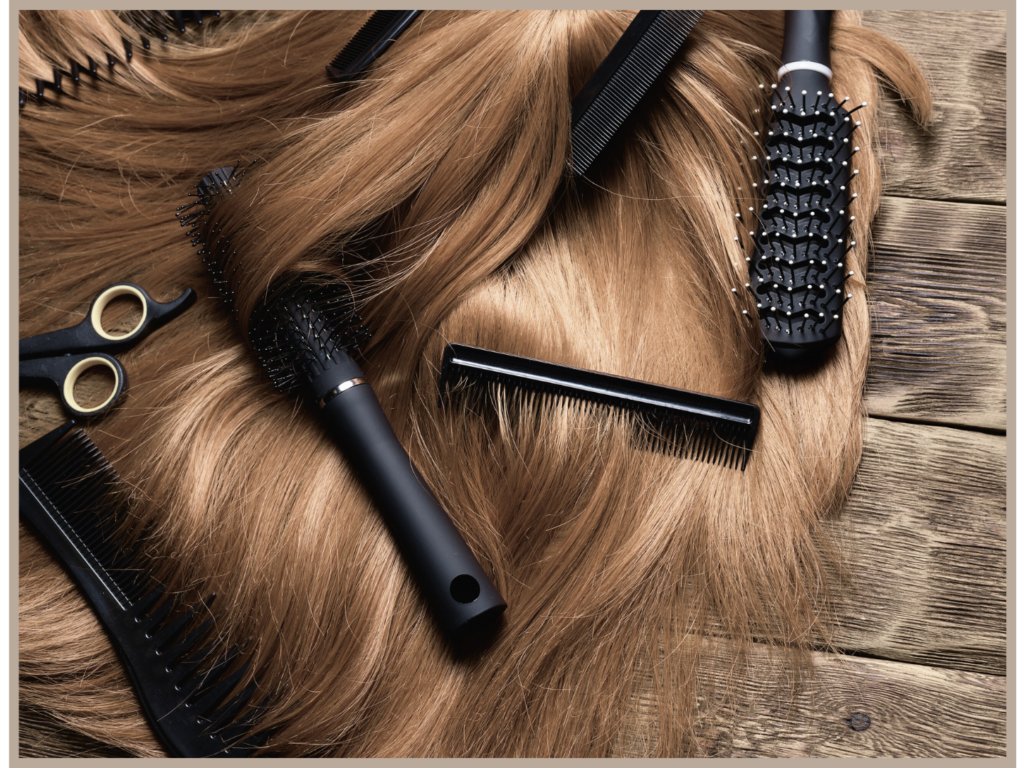 Hairbrushes, scissors, and a comb placed on a wooden surface around a section of long, blonde hair.
