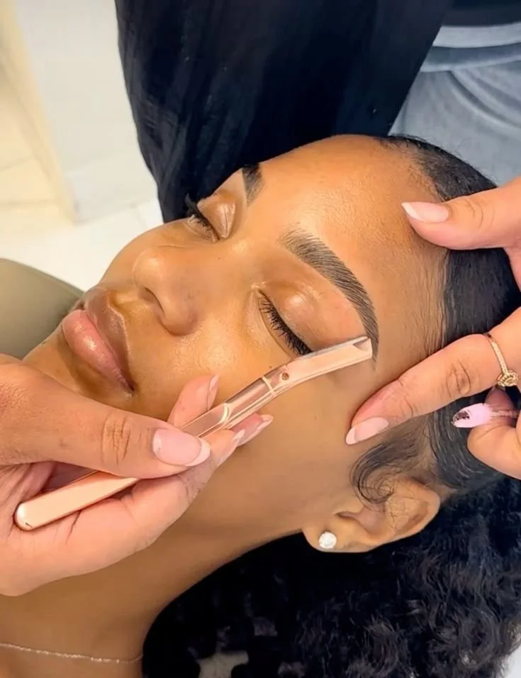 A woman in a beauty salon gets her eyebrows shaped with a wax applicator while her eyes are closed.