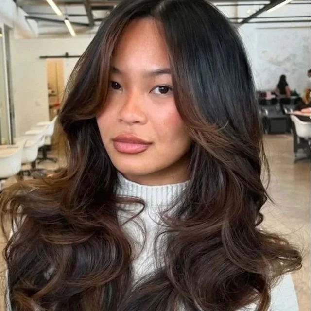 A woman with long, wavy brown hair and a white turtleneck top inside a modern, brightly lit space.